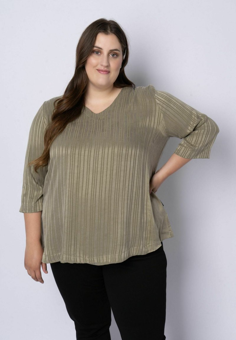 Femme aux longs cheveux bruns portant un haut côtelé vert olive à col en V et un pantalon noir, debout avec une main sur la hanche devant un fond uni.