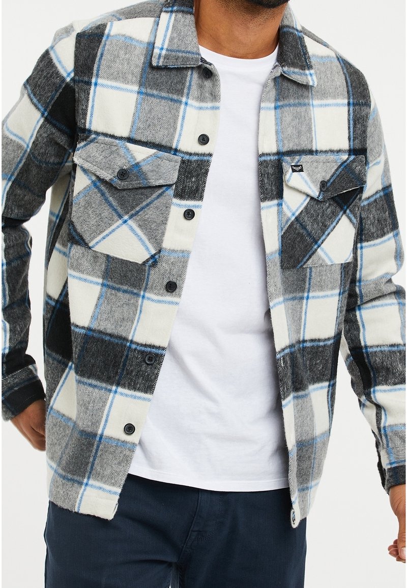 Homme portant une chemise à carreaux noire, grise, blanche et bleue boutonnée par-dessus un t-shirt blanc uni et un pantalon foncé, les mains près des hanches.