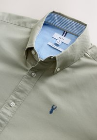Camicia verde chiaro con bottoni e colletto, caratterizzata da bottoni bianchi e un logo ricamato di un cervo blu. L'interno del colletto presenta righe blu.