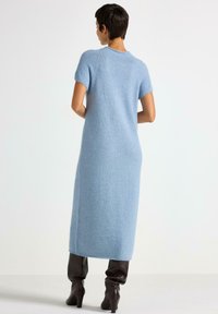 Nainen seisoo selkä kameraan päin, päällään vaaleansininen lyhythihainen neulemekko ja tummanruskeat korkokengät, jonka taustana on yksinkertainen valkoinen tausta.