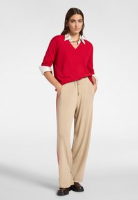 Nainen, joka pitää päällä punaista V-kaulaista neuletta valkoisen kauluspaidan päällä, beigejä leveälahkeisia housuja punaisilla raidalla sekä tummanruskeita kenkiä, seisoo kädet taskuissa.