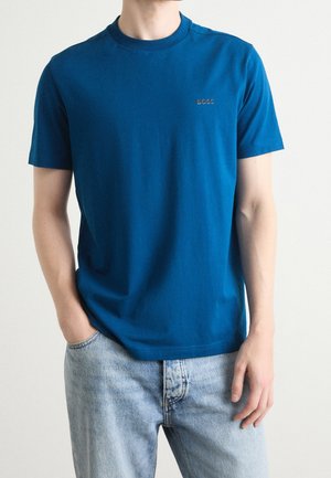 Mand iført en ensfarvet blå kortærmet t-shirt med "BOSS"-logo på brystet og lyseblå denimjeans, stående med den ene hånd i lommen.