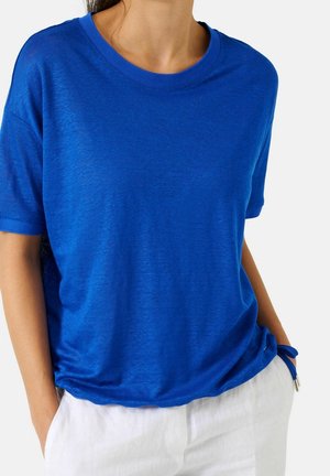 Persoon draagt een felblauw kortärmelig shirt met een ronde hals en een witte broek, handen casual in de zakken tegen een effen achtergrond.