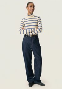 Mujer de pie con los brazos cruzados, vistiendo un cuello de tortuga a rayas en crema y azul, jeans de pierna ancha oscuros con cinturón y zapatos negros sobre un fondo claro.