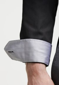 Polsino di camicia nera con una fodera a spina di pesce argentata, dotato di un bordo pulito e una sottile lavorazione. Il tessuto appare liscio e ben rifinito.