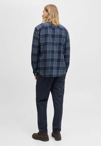 Camisa de franela de cuadros azul marino con mangas largas, que presenta líneas crema y azul claro, cierre de botones y un dobladillo curvado. Llevada con pantalones oscuros.
