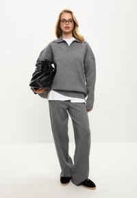Šedý pletený svetr s límečkem a oversized střihem, doplněný o volné šedé kalhoty ve stejném odstínu. Černá kabelka s nabíraným designem.