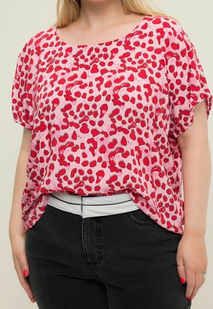 Kvinde iført løs lyserød top med røde abstrakte pletter, sorte jeans, hvidt bælte og rød neglelak på lys baggrund.