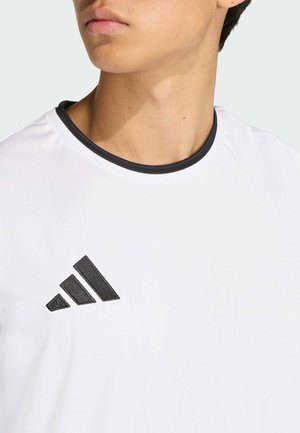 Młoda osoba w białej koszulce z czarnym okrągłym kołnierzem i czarnym logo z trzema paskami na górnej lewej części klatki piersiowej, lekko zwrócona na lewo.