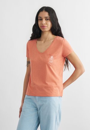 Młoda kobieta o długich ciemnych włosach, ubrana w koralowy T-shirt z dekoltem w serek i logo palmy na kieszeni oraz jasnoniebieskie spodnie w paski.