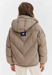 Osoba w beżowej pikowanej kurtce z kapturem, z kolorową naszywką na górnej części pleców, stojąca na tle białej ściany.