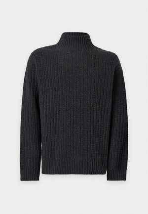 Homme portant un pull en maille à col roulé gris foncé, un jean bleu clair à jambes larges et des chaussures noires, debout devant un fond blanc uni.