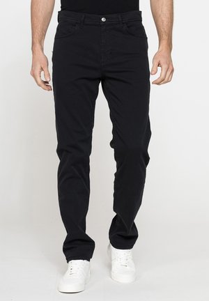 Uomo che indossa pantaloni neri a gamba dritta e scarpe da ginnastica bianche, in piedi davanti a uno sfondo chiaro e uniforme.