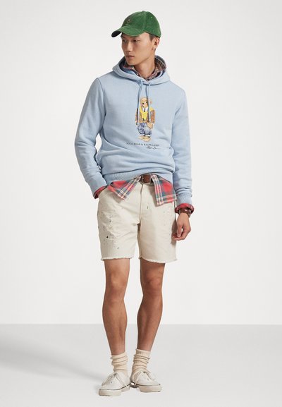 Γαλάζια hoodie με γραφικά σχέδια, φορεμένο πάνω από καρό πουκάμισο. Δεύτερες ρίγες σε εκρού σορτσάκι με φθαρμένο στρίφωμα και ελαφριές φθορές. Πράσινο καπέλο και μπεζ παπούτσια.