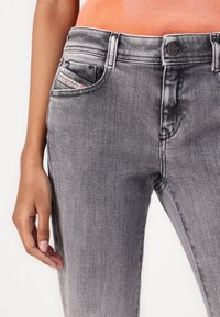 Detailní záběr osoby nosící šedé vybledlé džíny a oranžové tričko, ukazující levé uvolněné rameno podél těla a štítek značky Diesel na kapse.