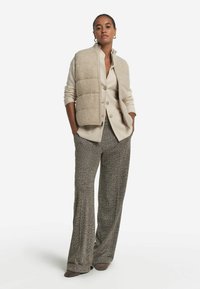 Beige pufferväst över en krämfärgad skjorta, kombinerad med bruna herringbonebyxor med vida ben och mörka skor. Släta material och skräddarsydd passform.