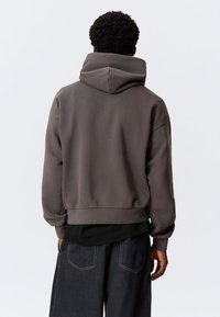 Mørk grå hettegenser med en stor hette, ribbestrikkede ermer og kant. Båret over en svart topp, sammen med mørke denimjeans. Glatt stofftekstur.