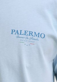 Ανοιχτό μπλε πουκάμισο με μπλε κείμενο "PALERMO Summer In Paradise SICILY ITALY" και μικρά χρώματα της ιταλικής σημαίας κάτω από το κείμενο.