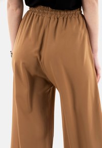 Pantaloni a gamba larga in un caldo tessuto marrone con una vita elastica. Texture liscia, senza modelli o accessori visibili, mostrati di spalle.