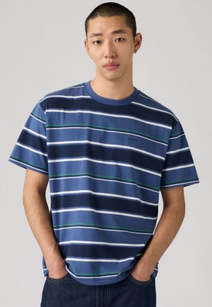 Młody mężczyzna z krótkimi włosami, ubrany w niebieską koszulę w paski z krótkim rękawem i ciemne spodnie, stoi z rękami w kieszeniach na jednolitym tle.