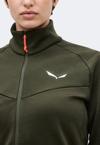 Κοντινή λήψη ατόμου που φορά ελιάς πράσινο μπουφάν με φερμουάρ και ψηλό γιακά, με μικρό λευκό λογότυπο στο στήθος, η ταινία του φερμουάρ είναι φωτεινό πορτοκαλί κορδόνι.