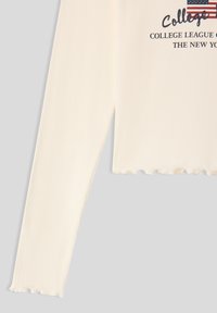 Top a maniche lunghe a coste color crema con orlo smerlato. Presenta una stampa grafica con testo e un design della bandiera americana.
