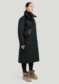 Lång svart vattentät kappa med hög krage, bärs över en svart polotröja och leggings, parat med tan isolerade stövlar och en svart axelremsväska.