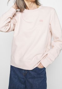 Felpa rosa chiaro con collo rotondo, polsini e orlo a coste. Presenta un piccolo logo ricamato sul petto. Abbinata a jeans in denim scuro.