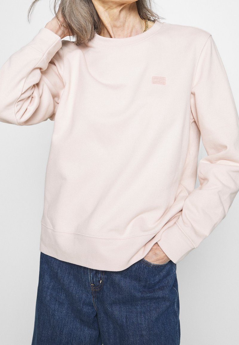 Felpa rosa chiaro con collo rotondo, polsini e orlo a coste. Presenta un piccolo logo ricamato sul petto. Abbinata a jeans in denim scuro.