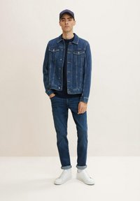Jachetă din denim slim-fit, de culoare albastru închis, cu buzunare frontale, purtată cu un pulover închis la culoare, blugi albastri și adidași albi. Bărbat stând pe un fundal deschis.