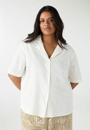 Donna che indossa una camicia bianca a maniche corte e bottoni, ampia, e pantaloni beige con un motivo a foglia bianca, in piedi contro uno sfondo bianco semplice.