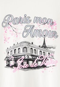 Hvid t-shirt med et sort og pink grafik af en pariserkafé med Eiffeltårnet, fremhævet af kirsebærblomst-motiver og kursiv skrift.