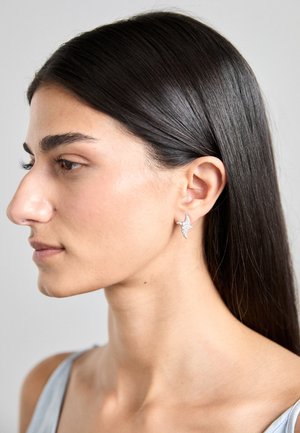 Boucles d'oreilles en argent en forme d'étoile, avec une surface texturée. Portées à l'oreille, avec de longs cheveux foncés lisses visibles.