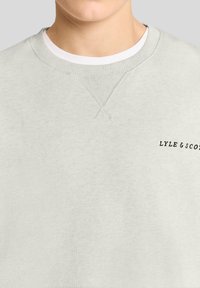 Κοντινό πλάνο ενός ατόμου που φοράει ανοιχτό γκρι φούτερ με λευκό μπλουζάκι με στρογγυλή λαιμόκοψη, με το κείμενο "LYLE & SCOTT" στο στήθος.