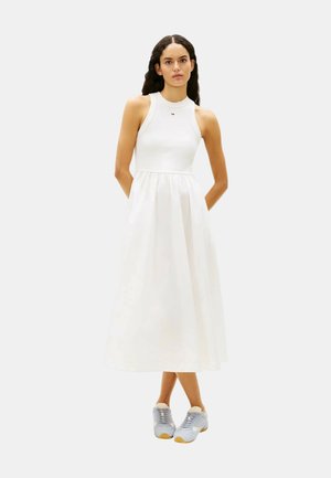 Kvinde med langt mørkt hår iført en ærmeløs hvid midi-kjole og lyseblå sneakers, stående med armene bag ryggen på ensfarvet baggrund.