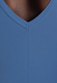 Gros plan d'une personne à la peau foncée portant un haut tricoté bleu à col en V, montrant la texture du tissu et le détail de l'encolure.