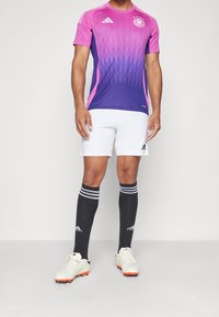 La camiseta de fútbol de Adidas presenta un diseño degradado que va del rosa al púrpura, con mangas cortas y un cuello redondo. Se combina con pantalones cortos blancos y calcetas negras.