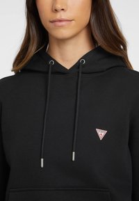 Felpa nera con cappuccio realizzata in materiale morbido, dotata di design a scollo a V, cordini con punte metalliche e un piccolo logo triangolare rosso e bianco sul petto.