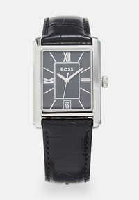 Montre carrée en argent avec un bracelet en cuir de crocodile noir. Le cadran noir présente des chiffres romains, des lignes verticales et un affichage de la date à 6 heures.