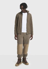 Beige Fleecejacke mit hohem Kragen und Reißverschluss, kombiniert mit strukturierten, beige Hosen mit einfarbigen, tanfarbenen Akzenten. Braune Wanderstiefel runden das Outfit ab.