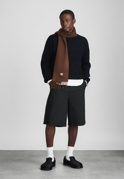 Maglione nero a maglia, sciarpa marrone con frange e logo, pantaloni corti neri, calze bianche e scarpe nere texture. Un outfit semplice e informale.