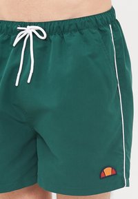 Zelené plavky s bielou šnúrkou, bočným lemovaním a kruhovým logom s oranžovými a žltými akcentmi. Hladká, ľahká tkanina.
