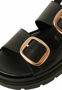 Black leather sandals feature two wide straps with a gold-tone buckle. The sole is thick and textured for stability, with a smooth inner lining.