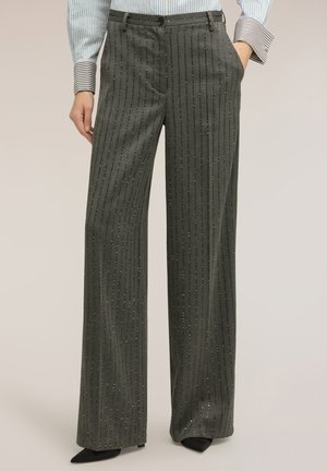 Osoba nosící šedé pruhované kalhoty s vysokým pasem a širokými nohavicemi s třpytivými detaily, ruce v kapsách, doplněné pruhovanou košilí a černými špičatými botami.