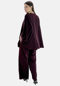 Weinrotes zweiteiliges Outfit mit fließendem Cape-Oberteil und weit geschnittenen Hosen. Hergestellt aus weichem Stoff mit glatter Textur und minimalen Nähten.