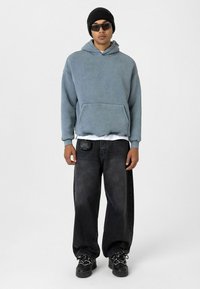 Lichtblauwe hoodie met een voorzak, gecombineerd met zwarte baggy jeans en sneakers. Accessoires zijn een zwarte beanie en een zonnebril.