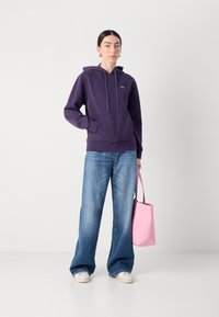 Violetiniai džemperiai su priekinėmis kišenėmis ir raišteliu, derinami su plačiomis mėlynomis džinsomis. Modelis laiko rožinę nešti maišą.