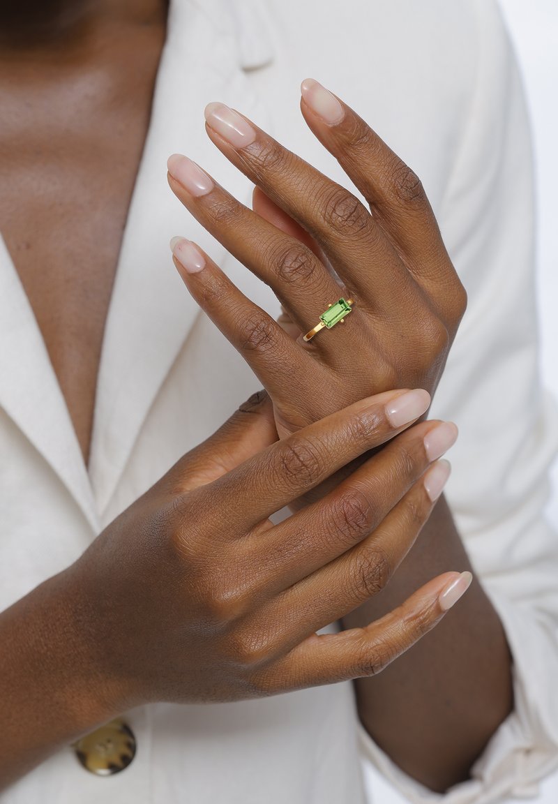 Gold ring with a rectangular green stone, worn on a slender finger. Hands have a smooth texture with neutral nail polish, against a light background.