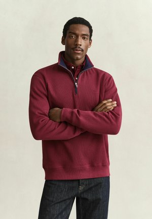 Man met gevouwen armen, die een bordeauxrode trui met een kwart ritssluiting draagt over een rood overhemd en donkerblauwe spijkerbroek, staande tegen een effen lichte achtergrond.