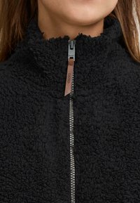 Svart fleecejakke med høy krage og glidelås. Teksturen ser myk og fluffy ut, med en kontrasterende glidelås og lærpulltab.